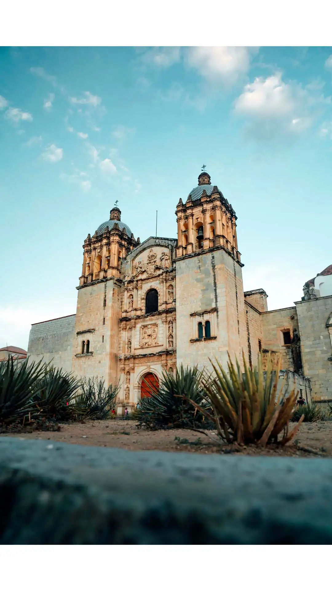 Templo de Santo Domingo de Guzmán en Oaxaca, con su arquitectura barroca y rodeado de plantas de agave bajo un cielo azul