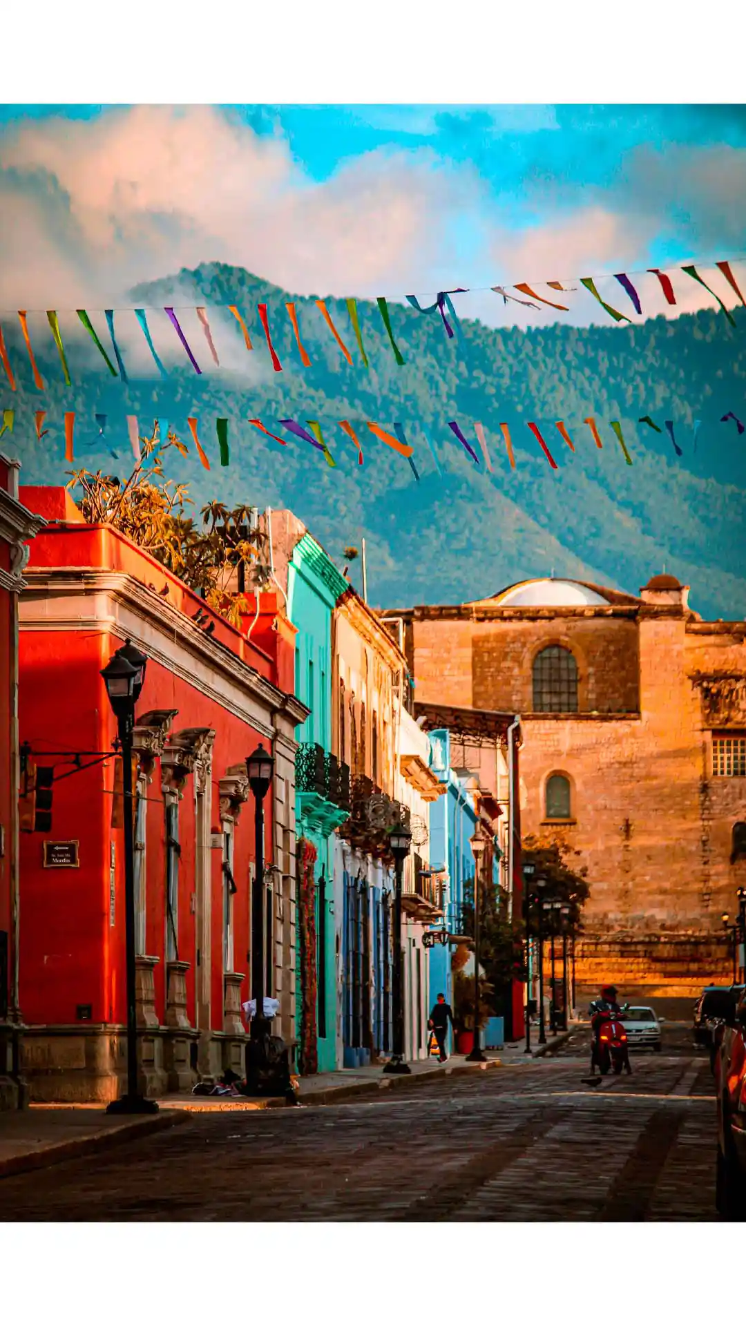 Calles coloridas del centro histórico de Oaxaca con montañas al fondo y banderines festivos sobre la avenida