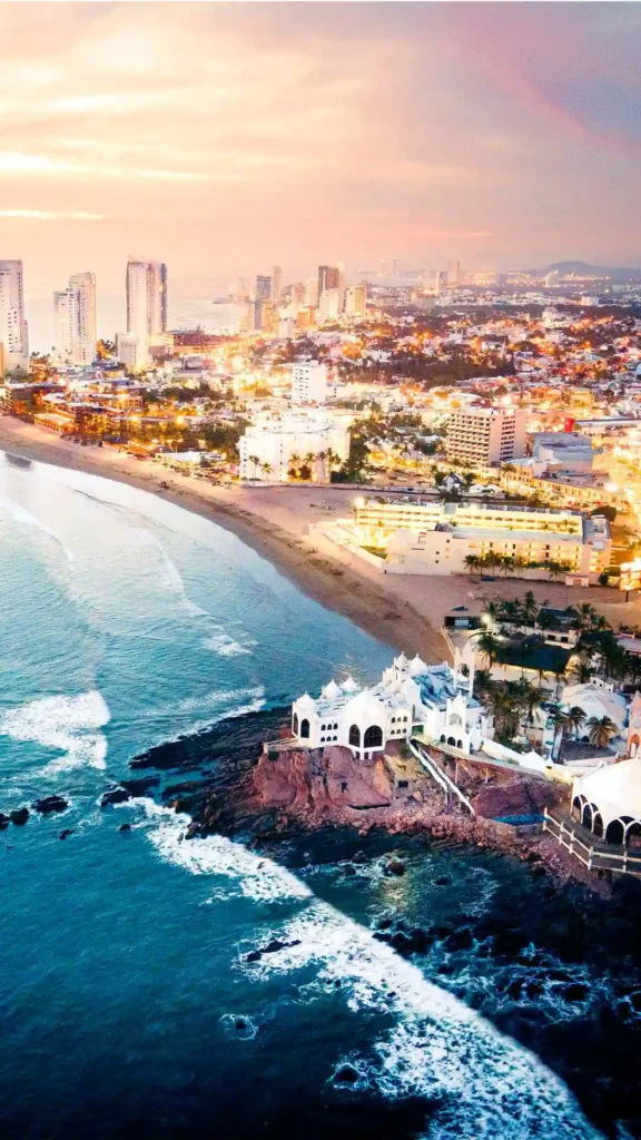 Vista aérea nocturna de Mazatlán iluminado junto al mar con edificios, playa y olas del Pacífico