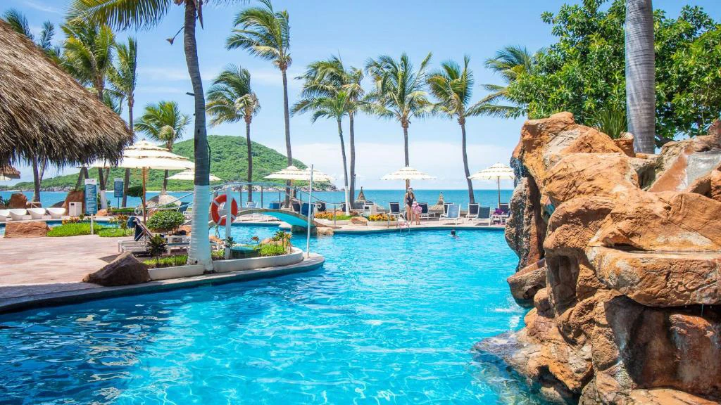 Piscina del Hotel Luna Palace Mazatlán con palmeras, camastros y vista al mar en un entorno tropical.