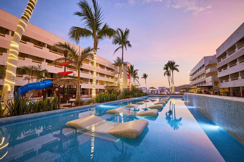 Vista al atardecer de la piscina principal del Hotel Emporio Mazatlán con palmeras y áreas recreativas.